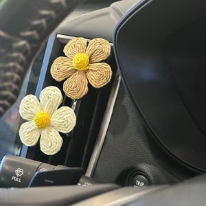 May include: Two daisy-shaped car air fresheners. One is cream-coloured with a yellow centre, the other is tan with a yellow centre. The air fresheners are made of woven material and are clipped into a car's air vent.