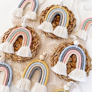 May include: A collection of handmade rainbow wall hangings with a macrame design. Each rainbow features a different pastel colour scheme, with white fringe and wooden bead accents. The rainbows are displayed on woven circular placemats.
