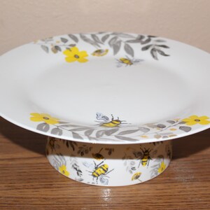 May include: A white ceramic cake stand featuring a pattern of yellow flowers, gray leaves, and bumblebees. The circular plate has a wide rim, and the cylindrical base is decorated with a matching botanical and insect motif in yellow and gray.