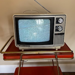 May include: A vintage AMGA Gold Star television with a silver antenna and control knobs sits on a red serving cart with brass-colored handles. The TV screen displays static. The cart has two red trays.