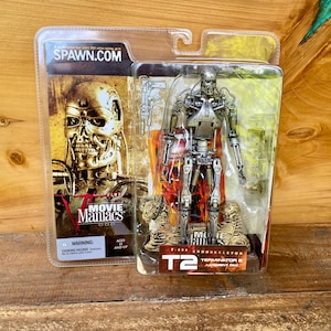 May include: A packaged T-800 Endoskeleton action figure from the Terminator 2: Judgment Day movie. The figure is silver and has two guns. The packaging includes the text "SPAWN.com" and "Movie Maniacs".