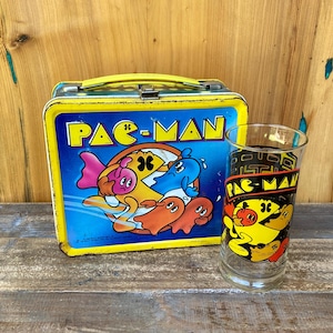 May include: Vintage Pac-Man lunchbox and glass. The lunchbox is yellow and blue with a Pac-Man graphic. The glass features Pac-Man characters. Both items are retro and nostalgic, perfect for fans of the classic arcade game.
