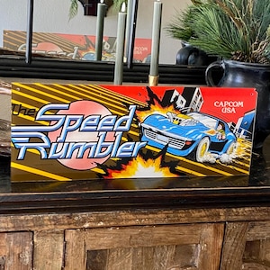 May include: A vintage arcade game sign for "The Speed Rumbler" featuring a blue sports car racing. The sign has a red and yellow background with the Capcom USA logo. The text "The Speed Rumbler" is in blue and white.