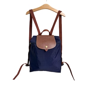 May include: A navy blue backpack featuring a brown leather flap with a gold-tone snap button closure. The bag includes a top handle and two adjustable brown fabric shoulder straps. The front flap displays an embossed logo and a textured finish.