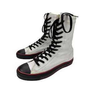 May include: A pair of white high-top sneakers featuring black laces, black rubber toe caps, and black soles with a thin red stripe detail. These tall lace-up shoes have silver-toned metal eyelets and a structured ankle-high design.