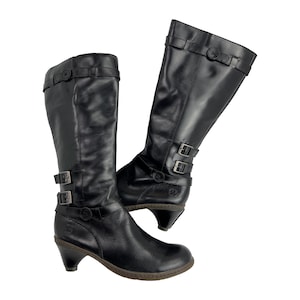 May include: A pair of black leather knee-high boots. Each boot features a rounded toe, a small heel, and adjustable straps with silver-tone buckles. The boots have a classic, slightly distressed appearance.