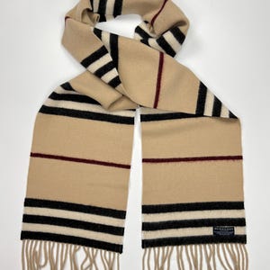 May include: A beige scarf with black and white stripes and a burgundy stripe. The scarf has a fringed edge and a small label that says "Burberry". The scarf is folded and draped.