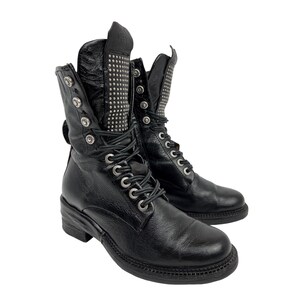 May include: Black leather combat boots with silver-tone hardware. The boots feature a lace-up design, multiple buckles, and a studded panel on the upper shaft. The soles are black and have a low heel.