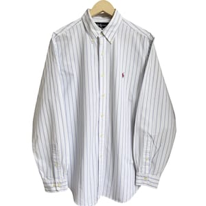 Può includere: Camicia bianca con bottoni e righe verticali blu. La camicia a maniche lunghe presenta un colletto button-down e un piccolo logo rosso sul petto. La camicia è appesa a una gruccia di legno.