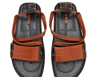 May include: Leather sandals in brown and orange. The sandals have a strap design, with the word "PRADA" visible on the straps. The soles are dark grey with white stitching. The sandals are suitable for warm weather.