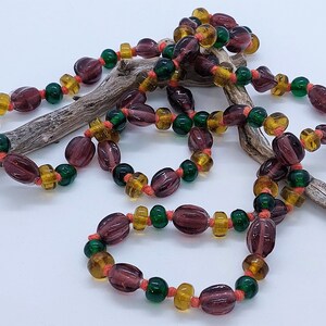 May include: A long necklace made of purple, green, and orange glass beads. The beads are strung on a thin cord and are arranged in a repeating pattern.