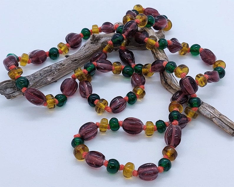 May include: A long necklace made of purple, green, and orange glass beads. The beads are strung on a thin cord and are arranged in a repeating pattern.