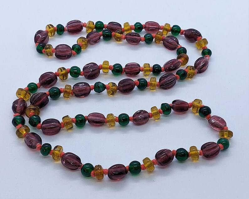 May include: A long necklace made of purple, green, and orange glass beads. The beads are strung on a thin cord and are arranged in a repeating pattern.