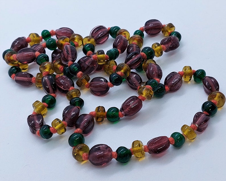 May include: A long necklace made of purple, amber, and green glass beads. The beads are strung on a thin orange cord.