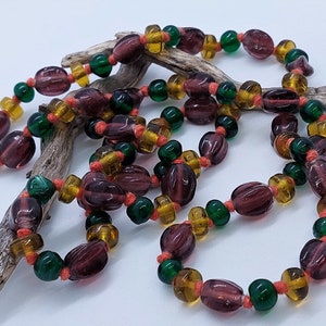 May include: A long necklace made of glass beads in shades of purple, green, and amber. The beads are strung on a thin cord and are tied together with small knots.