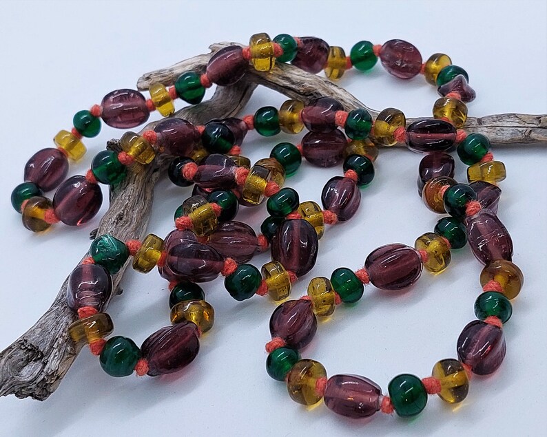 May include: A long necklace made of glass beads in shades of purple, green, and amber. The beads are strung on a thin cord and are tied together with small knots.