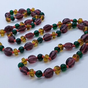 May include: A long necklace made of purple, green, and orange glass beads. The beads are strung on a thin cord and are arranged in a repeating pattern.