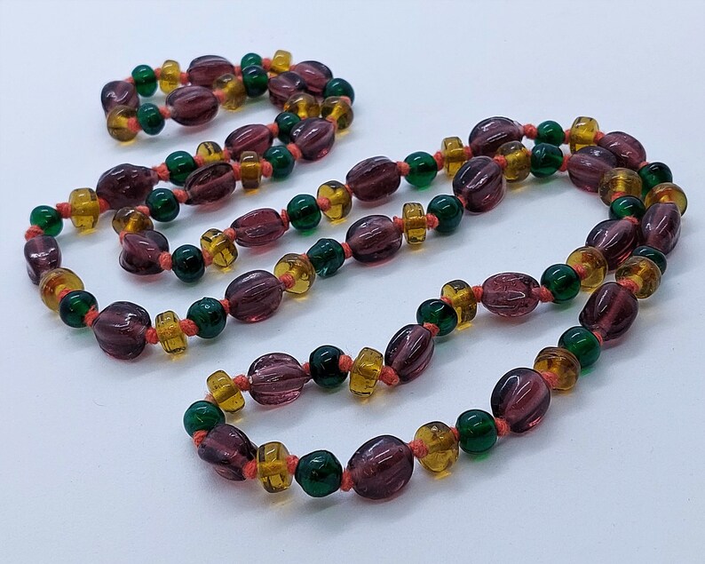 May include: A long necklace made of purple, green, and orange glass beads. The beads are strung on a thin cord and are arranged in a repeating pattern.