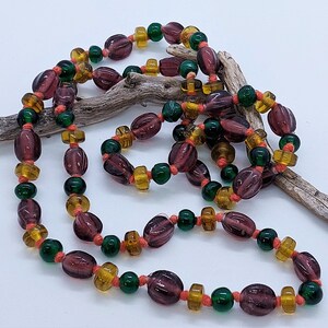 May include: A long necklace made of purple, green, and amber glass beads. The beads are strung on a thin cord and are arranged in a repeating pattern.