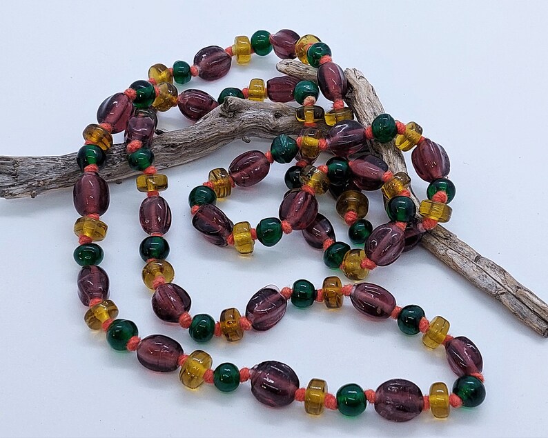 May include: A long necklace made of purple, green, and amber glass beads. The beads are strung on a thin cord and are arranged in a repeating pattern.