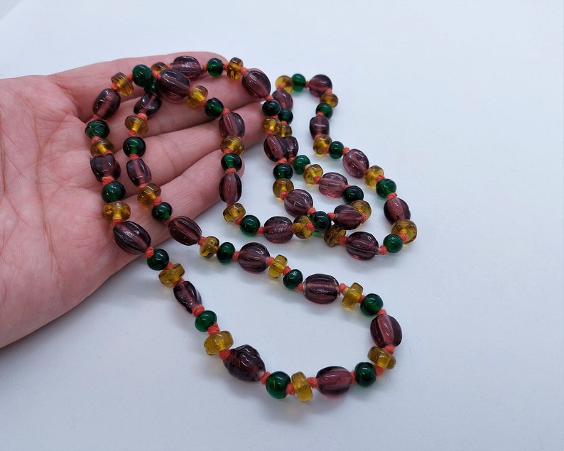 May include: A long necklace made of purple, green, and orange glass beads. The beads are oval shaped and have a glossy finish.