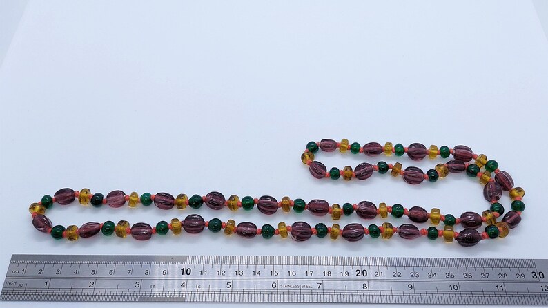 May include: A long necklace made of glass beads in shades of purple, orange, and green. The beads are strung on a clear cord and are approximately 30 inches long.