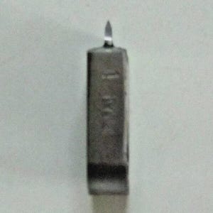 May include: A close-up of a small, rectangular, metallic object with a pointed tip. The object appears to be a tool or instrument, possibly for precision work. The surface has some markings, but they are not clear.