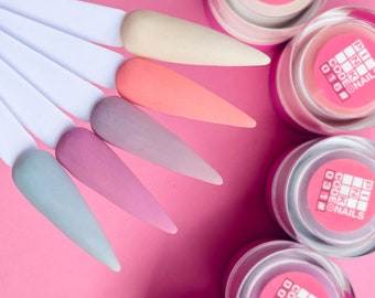 Puede incluir: Cinco pequeños frascos de polvo acrílico para nail art. Los frascos están etiquetados con el texto "CODE" y un número. Los colores del polvo son beige, coral, rosa claro, lavanda y azul claro.