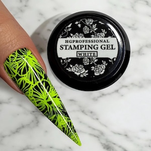 May include: A small black jar with a white label that says "HGPROFESSIONAL STAMPING GEL WHITE" and a floral design. The jar is on a white marble surface. A fingernail with a black and neon green nail design is visible in the background.
