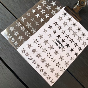 May include: Two sheets of nail art stickers with different star designs. One sheet has white stars on a clear background. The other sheet has black stars and geometric star designs on a white background. The text on the sheet reads "HGPROF.COM STARS-3357".
