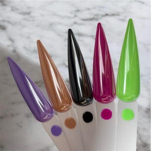 May include: Five long, pointed nail tips in a row, each with a different glossy color: purple, brown, black, magenta, and green. Each nail is mounted on a white stick with a matching colored dot at the base. The background is a marble pattern.