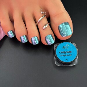 May include: Close-up of a foot with chrome cerulean nail polish. The toenails are painted with a shiny, iridescent teal color. A silver snake toe ring is worn on the second toe. A small jar of HG Chrome Cerulean nail powder is next to the foot.
