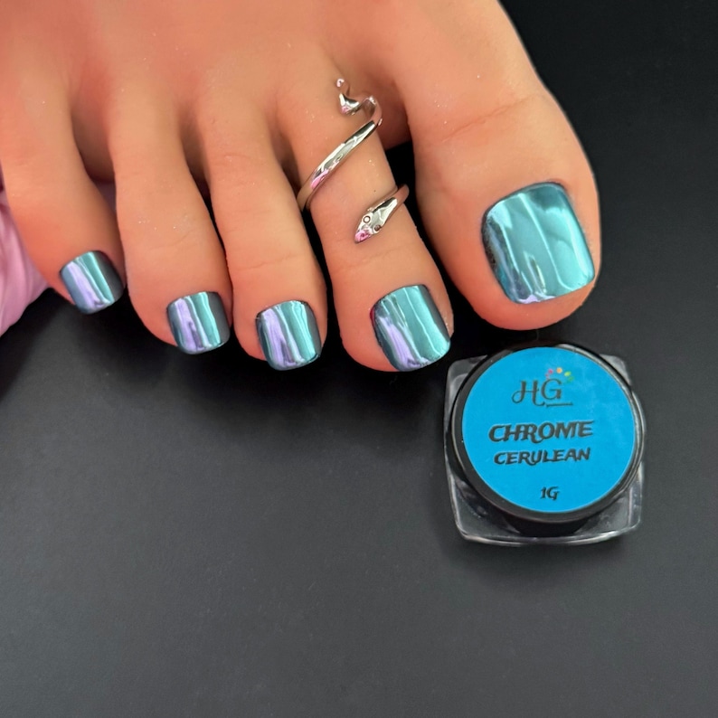 May include: Close-up of a foot with chrome cerulean nail polish. The toenails are painted with a shiny, iridescent teal color. A silver snake toe ring is worn on the second toe. A small jar of HG Chrome Cerulean nail powder is next to the foot.