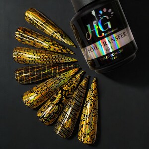 Op de afbeelding: Een fles HG Professional Foil Transfer Gel, een heldere gel met een zwart etiket en zilveren letters. De fles is omringd door tien nageltips met verschillende gouden folieontwerpen.
