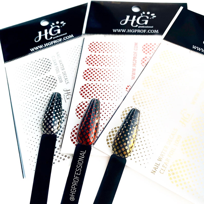 Puede incluir: Tres hojas de calcoman&iacute;as de agua para u&ntilde;as con un patr&oacute;n de estrellas. Las calcoman&iacute;as son de color dorado, rojo y plateado. Las hojas est&aacute;n etiquetadas con el nombre de la marca "HG" y el sitio web "www.hgprof.com".