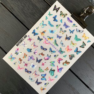 Puede incluir: Una hoja de calcoman&iacute;as para u&ntilde;as con forma de mariposa de colores sobre un fondo blanco. Las calcoman&iacute;as presentan mariposas en varios tonos de azul, rosa, morado, verde y amarillo. La hoja tambi&eacute;n incluye el texto "BUTTERFLY_002" y "WWW.HGPROF.COM".