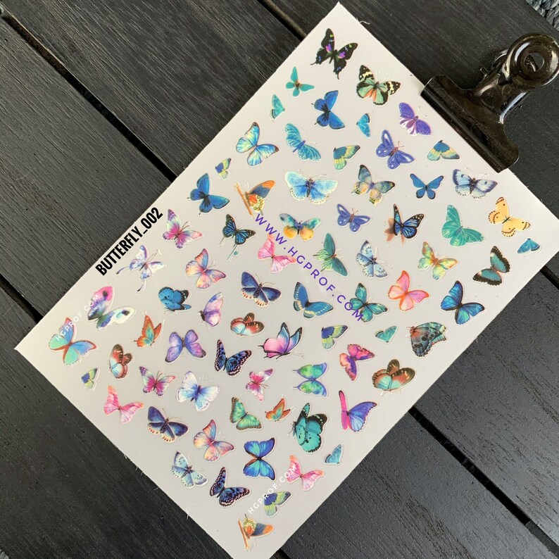 Puede incluir: Una hoja de calcoman&iacute;as para u&ntilde;as con forma de mariposa de colores sobre un fondo blanco. Las calcoman&iacute;as presentan mariposas en varios tonos de azul, rosa, morado, verde y amarillo. La hoja tambi&eacute;n incluye el texto "BUTTERFLY_002" y "WWW.HGPROF.COM".