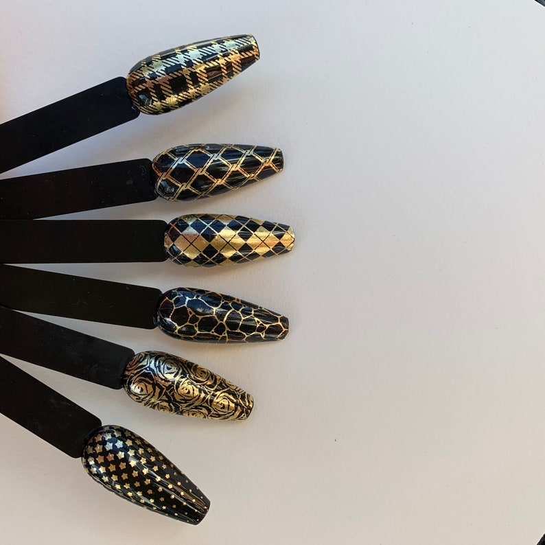 Puede incluir: Seis tiras de esmalte de u&ntilde;as negras y doradas con diferentes patrones geom&eacute;tricos. Las tiras est&aacute;n dispuestas en forma de abanico sobre una superficie blanca.