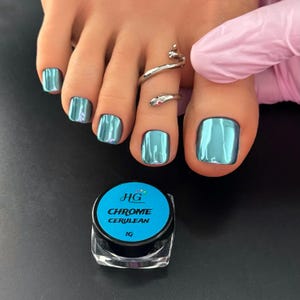 May include: A close-up of a foot with teal chrome nail polish and a silver snake toe ring. A small jar of "HG Chrome Cerulean" nail polish is in the foreground. The nails have a shiny, metallic finish.