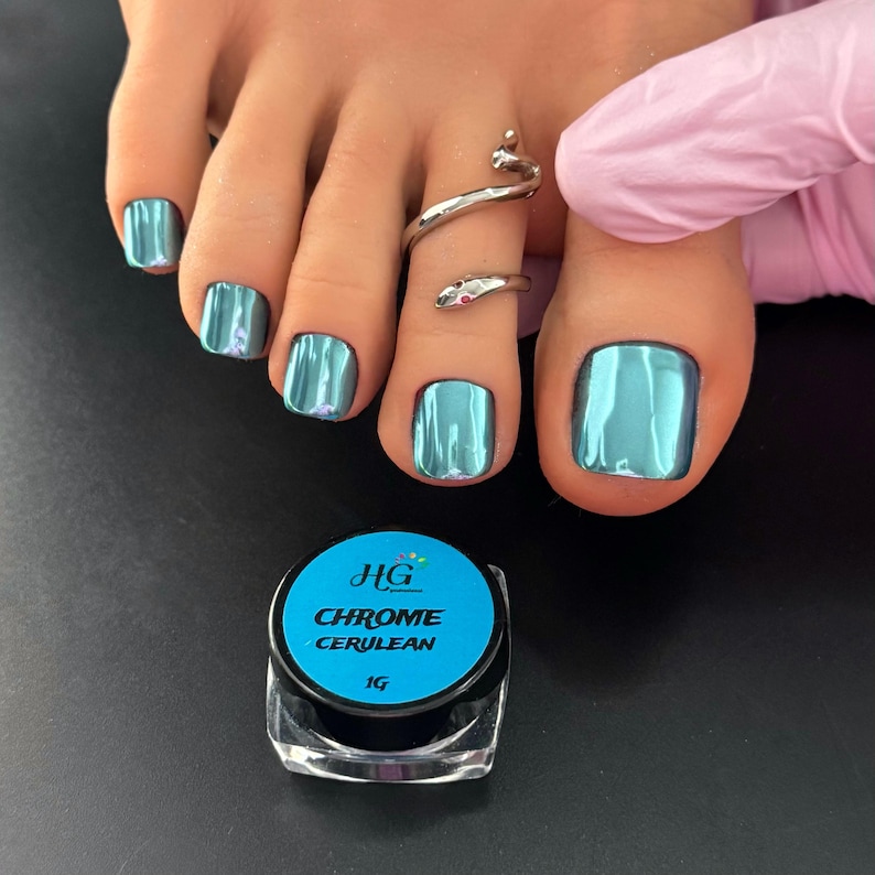 May include: A close-up of a foot with teal chrome nail polish and a silver snake toe ring. A small jar of "HG Chrome Cerulean" nail polish is in the foreground. The nails have a shiny, metallic finish.