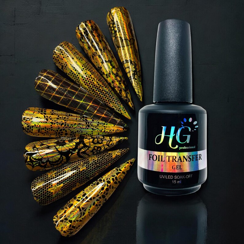 Op de afbeelding: Een flesje HG Professional Foil Transfer Gel, een heldere gel met een zwart etiket en gouden letters. Het etiket vermeldt "HG Professional Foil Transfer Gel UVLED Soak-Off 15 ml". De fles wordt omringd door verschillende nageltips met verschillende gouden folieontwerpen.