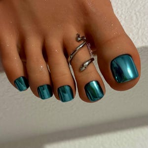 May include: Close-up of a foot with teal metallic nail polish. A silver snake toe ring wraps around the second toe. The nails have a glossy, reflective finish, and the skin appears moisturized.
