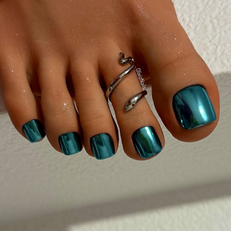 May include: Close-up of a foot with teal metallic nail polish. A silver snake toe ring wraps around the second toe. The nails have a glossy, reflective finish, and the skin appears moisturized.