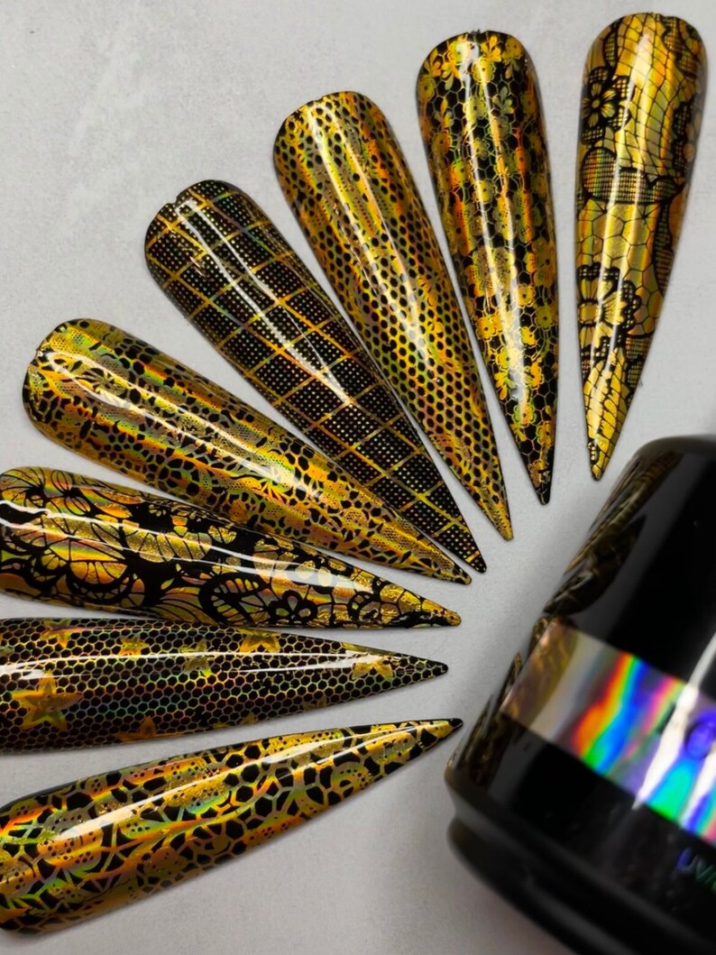 Op de afbeelding: Een set van tien gouden en zwarte nagelfolies met verschillende geometrische en bloemmotieven. De folies zijn in een waaier op een wit oppervlak gerangschikt. Een zwarte fles met een regenboogkleurig, iriserend etiket staat op de achtergrond.