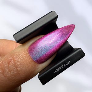 Puede incluir: Primer plano de una uña con un vibrante esmalte iridiscente rosa y azul. La uña está sujeta por una herramienta de nail art negra y texturizada. El sitio web HGPROF.COM es visible.