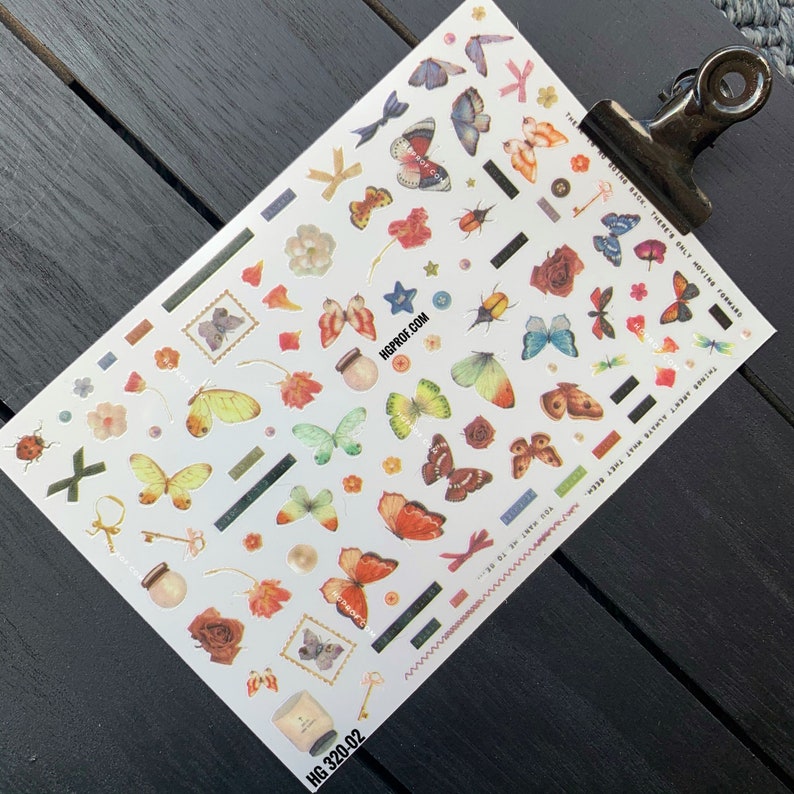 Puede incluir: Una hoja de pegatinas de mariposas de colores con una variedad de dise&ntilde;os, incluyendo flores, estrellas, llaves y un frasco con una mariposa dentro. La hoja es blanca con un borde negro y el texto "HGPROF.COM" y "HG 320-02" impreso en ella.
