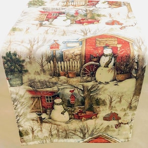 May include: A white table runner with a winter scene featuring snowmen, red barns, and trees. The background is a light beige with a subtle sparkle.