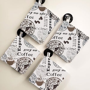 May include: Four white square coasters with a coffee theme. The coasters feature a brown and white design with coffee cups, coffee beans, and the words "Latte", "Espresso", "Cappuccino", and "Coffee".