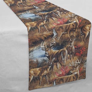 May include: A rectangular table runner featuring a detailed print of deer in a natural landscape. The design includes various deer poses, a stream, and autumnal foliage in shades of brown, tan, and red. The runner is displayed on a white surface.