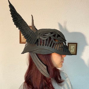 May include: A black helmet with a winged design and a detailed, intricate pattern. The helmet has a long, pointed horn on the top and a wide brim.
